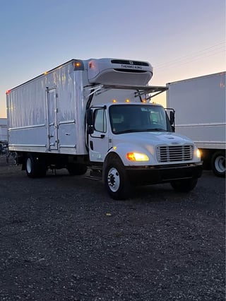 White refrigerated box truck with Thermo King unit parked in lot at dusk, headlights on