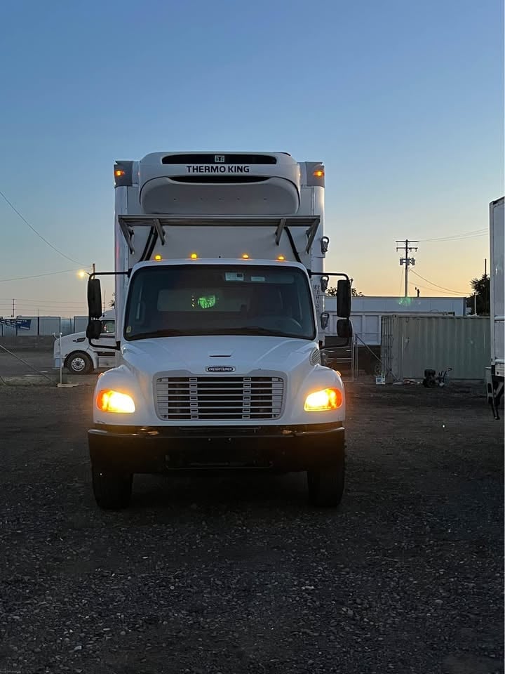 White refrigerated delivery truck with Thermo King unit parked in a lot at dusk with headlights on