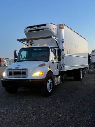 White Freightliner refrigerated truck with Thermo King unit parked in a lot, headlights on against blue sky
