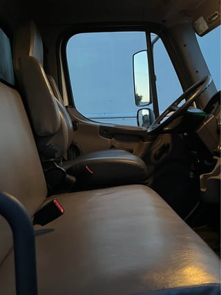 Interior view of a truck cab with tan seats, steering wheel, and windshield showing clear blue sky