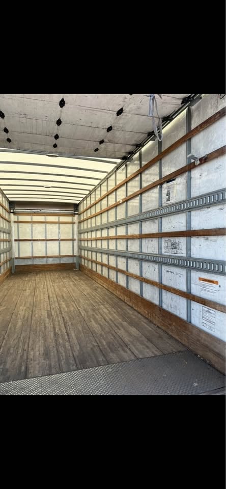 Interior of an empty cargo truck bed with wooden flooring, metal walls, and roof lights visible from the open rear door