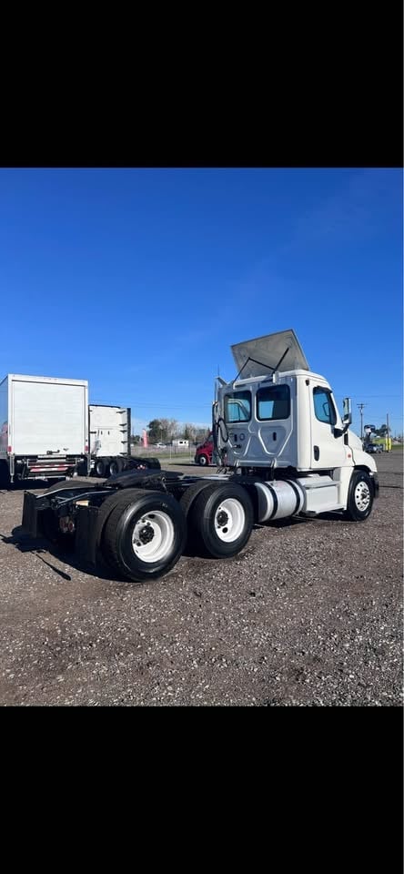White semi-truck with gray cab on asphalt lot, other commercial vehicles visible in background against clear blue sky