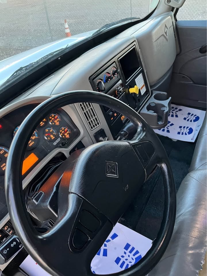Interior of a truck cab with black steering wheel, dashboard with illuminated orange gauges, and blue papers on the seats