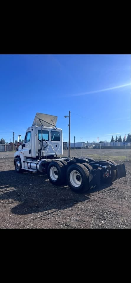 White semi-truck with open hood parked in lot under blue sky with evergreen trees visible in background