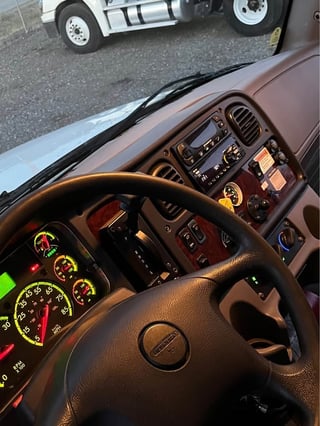 Interior view of a semi truck cab showing steering wheel, dashboard gauges, and controls with another truck visible outside