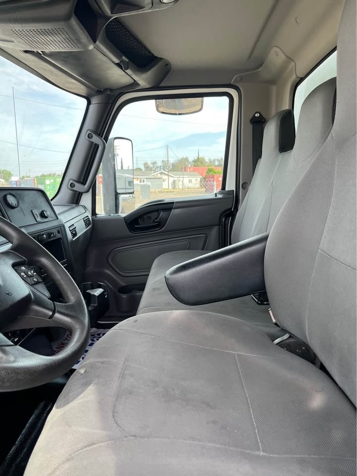 Interior of a truck cab showing steering wheel, gray seats, and windshield view of a parking lot