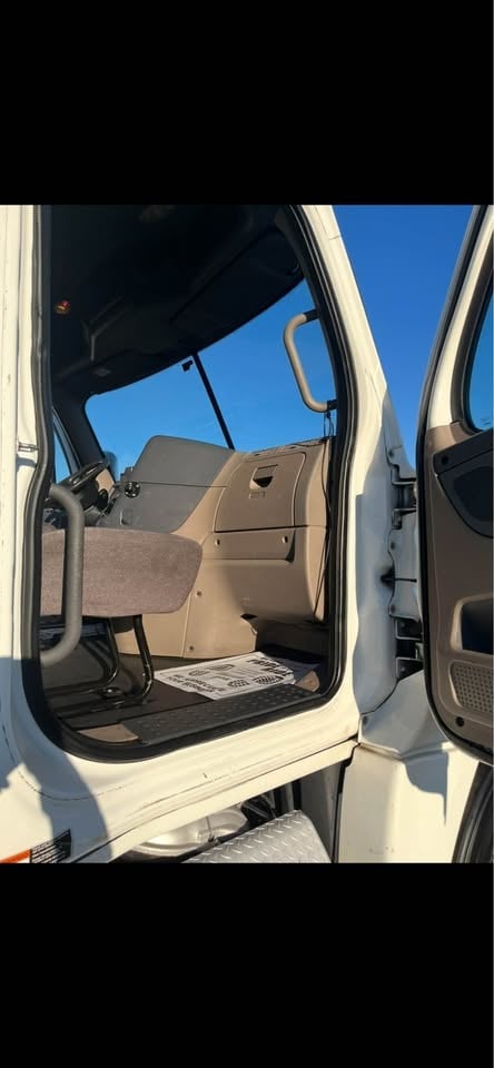 Interior view of a white truck cab with open door, showing tan seats, steering wheel, and clear blue sky visible through windshield